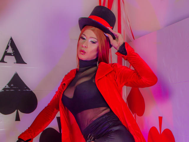 EsmeraldaLipss porte un manteau rouge, un haut noir transparent et un pantalon en cuir noir, avec un chapeau haut noir, qu'elle tient, montrant ses seins et son rouge à lèvres rouge, dans une pose sexy..