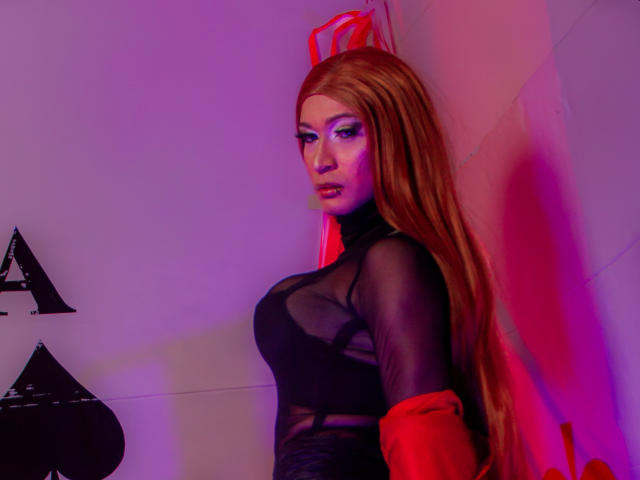 Esmeralda Lipss mit langen, roten Haaren, die ein schwarzes, durchsichtiges Oberteil, rote Handschuhe und pinkes Augen-Make-up trägt, steht in violett-rotem Licht und zeigt ihre Brüste..