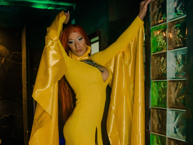 Esmeralda Lipss mit langen, roten Haaren, einem gelben Latekleid und freiliegenden, großen Brüsten, mit erhobenen Armen, stehend in einem dunklen Raum mit grünem Licht, mit Glasblöcken auf der rechten Seite..