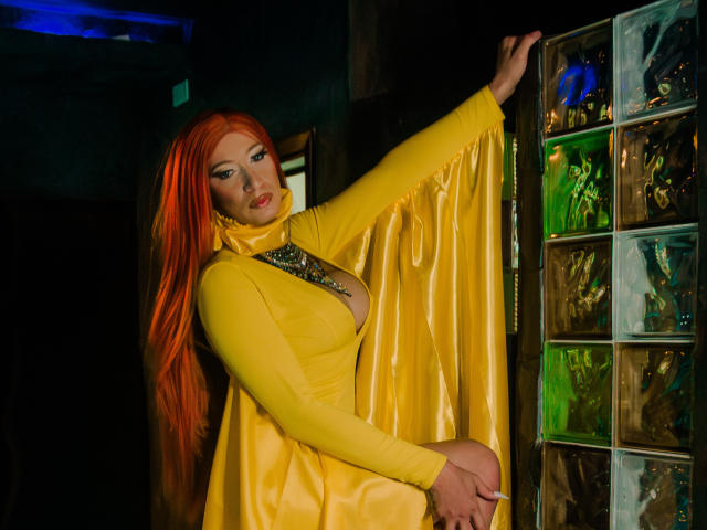 EsmeraldaLipss met feloranje haar, gekleed in een gele satijnen jurk met een diepe decolleté, die een glazen muur aanraakt en verleidelijk naar de camera kijkt..
