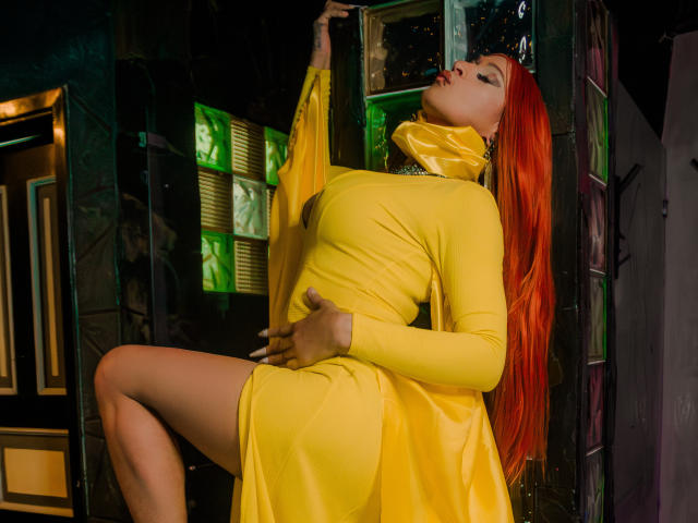 Esmeralda, con i capelli rosso acceso, un vestito giallo e una sciarpa gialla lucente, appoggiata a un muro scuro, con una mano sulla coscia, una gamba sollevata, gli occhi chiusi e le labbra leggermente dischiuse..