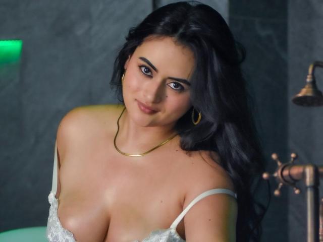 AmberGatess tiene senos grandes y desnudos, vistiendo un sujetador blanco de encaje, con cabello negro largo, aros de oro y un collar de oro, sonriendo ligeramente. Está en una habitación oscura..