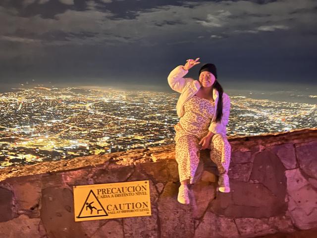 Colette Bessette, com longos cabelos pretos, vestindo uma jaqueta branca e calças de estampa de leopardo, sentada em um degrau de pedra, apontando para cima, luzes da cidade abaixo, céu noturno escuro e nublado..