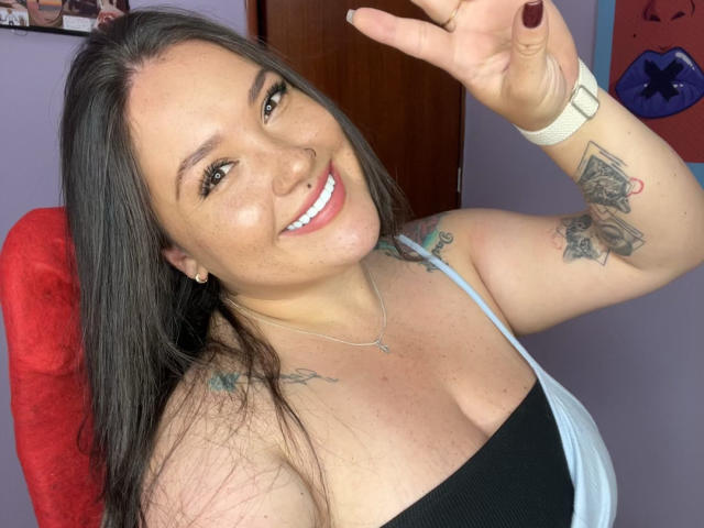 Colette Bessette, con i capelli scuri, sorridente, indossa una maglietta nera e blu, che mostra il seno, ha tatuaggi sul braccio e sul petto, labbra rosa, un braccialetto bianco e alza la mano destra..