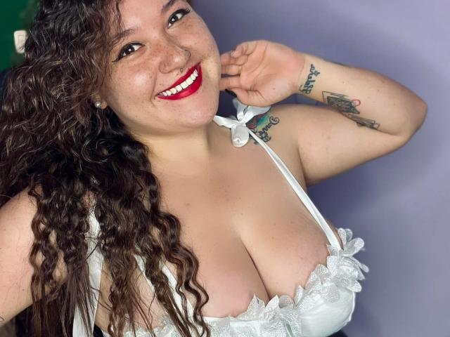 Colette Bessette, com cabelos castanhos cacheados, sorrindo, batom vermelho, sutiã de renda branco, seios grandes, tatuagem no braço direito, segurando o queixo, fundo de parede branco..