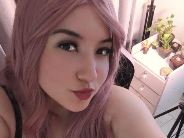 SusanMonroy with pink hair, black eyeliner, red lipstick, and a cute smile, showing her bare shoulders, wearing a black top, sitting near a white dresser with a plant.