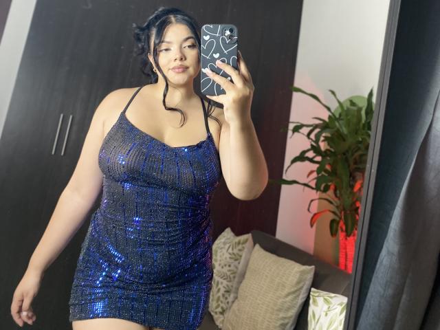 LaraJames, con i capelli neri e un abito blu ricoperto di paillettes, tiene in mano un telefono con una custodia bianca a motivi, scattando un selfie allo specchio, mostrando il suo seno prosperoso e i glutei..