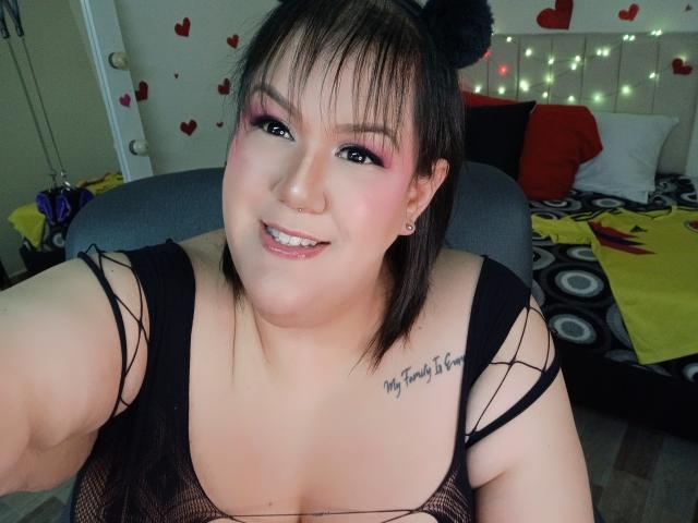 GiiannaMorrys, avec un haut en résille noire, du fard à paupières rose, des cheveux noirs attachés en chignon, souriante, montrant de petites seins, un tatouage sur l'épaule droite, dans une chambre avec un lit et des autocollants en forme de cœur..