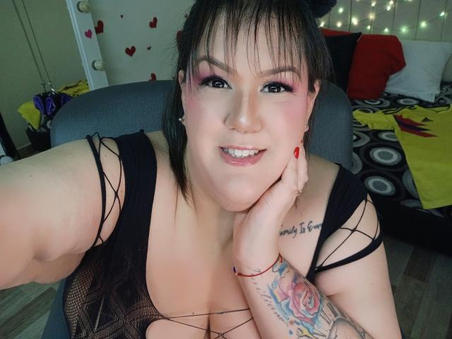 GiiannaMorrys with pink eye makeup, black mesh top, large tits, red nail polish, tattooed arm, hand on cheek, smiling, black hair, bedroom, string lights in background.