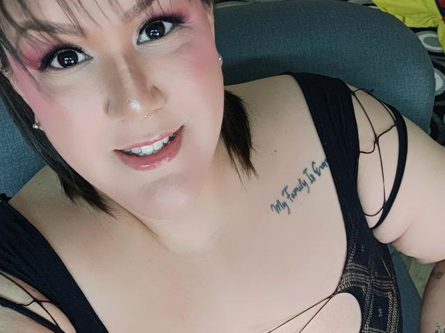 GiiannaMorrys has pink eyeshadow, a nose piercing, and a tattoo on her chest. She's wearing a black, strappy top, showing her tits, and smiling at the camera.