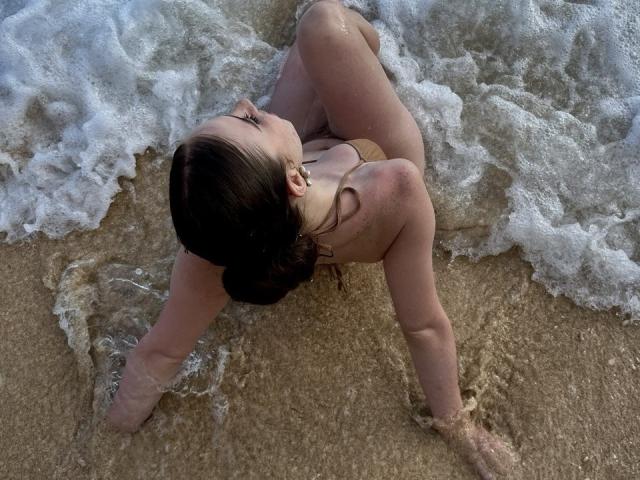 FelicitasDeroy is naakt, zittend op nat zand met golven om haar heen. Haar achterkant is zichtbaar, en ze heeft donkerbruin haar, kijkt omhoog met gespreide armen..