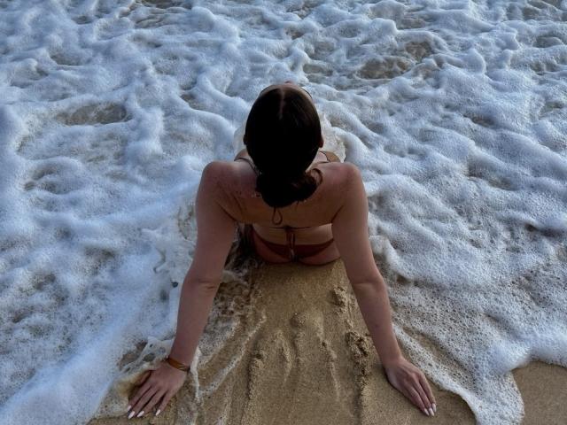 FelicitasDeroy, face à la caméra, assise dans le sable mouillé avec des vagues mousseuses, portant un bikini rouge, bras ouverts, cheveux bruns attachés en chignon, marques de bronzage, bracelet au poignet droit..
