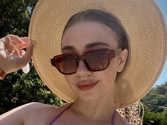 Felicitas Deroy draagt een grote, bruine zonnehoed, bruine zonnebril, een neusring en roze lippenstift, en laat haar schattige gezicht zien met een lichte glimlach. Ze draagt een paars bikini top..