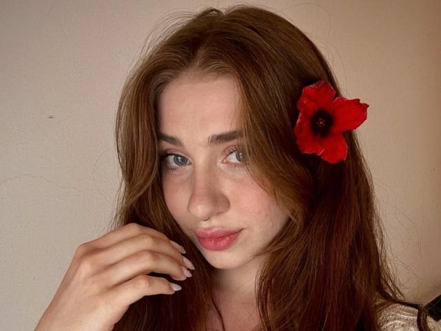 Felicitas Deroy tiene el pelo largo y castaño, una flor roja en el pelo, piel clara, ojos azules y labios rosados ligeramente separados. Está tocándose la barbilla con la mano derecha..