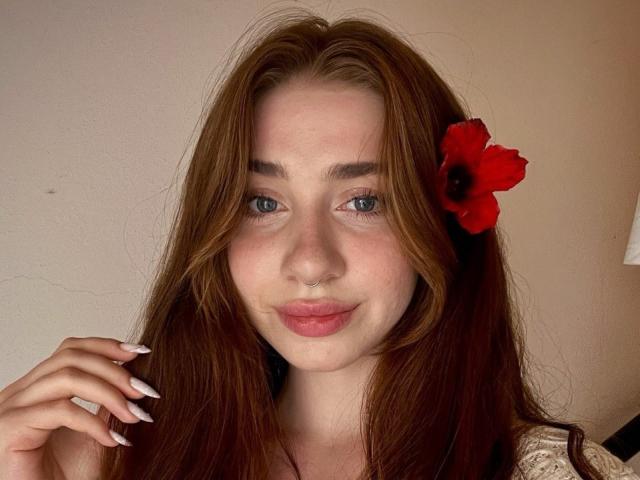Felicitas Deroy, met lang rood haar, blauwe ogen, een neusring en een rode bloem in haar haar, lachend met roze lippen, haar haar vasthoudend, met witte nagels, lichte huid en een schattig gezicht..