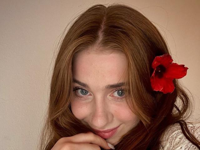 FelicitasDeroy a les cheveux roux, une fleur rouge dans ses cheveux, et des yeux bleus, avec un sourire discret et sa main près de sa bouche. Ses seins ne sont pas visibles..