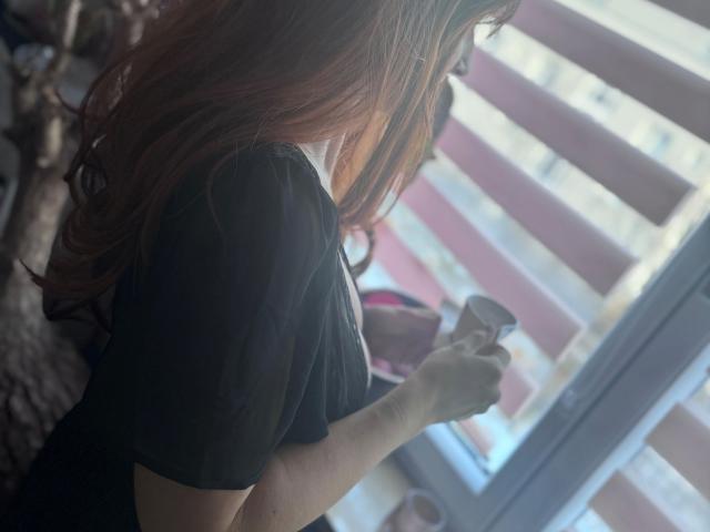 BellaMila-hot with long red hair, wearing a black shirt, holding a glass in her right hand, standing by a window with blinds.