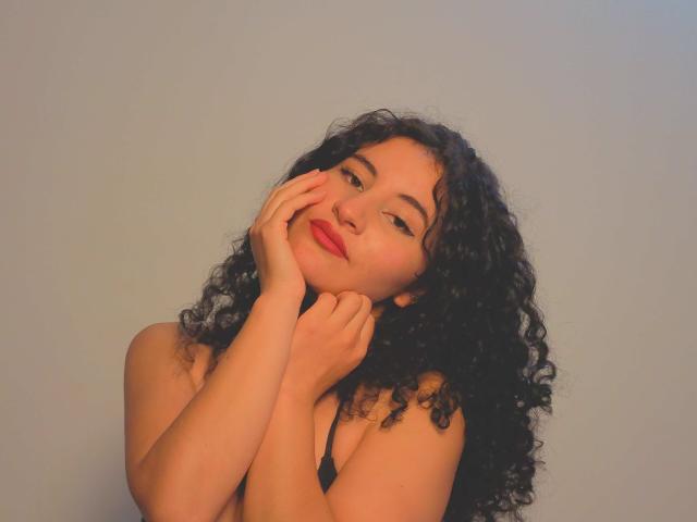 DesireLoah, con cabello negro rizado, labios rojos y manos que suavemente tocan su rostro, mirando hacia arriba con una sonrisa delicada, vestida con una correa negra, sobre un fondo gris claro liso..