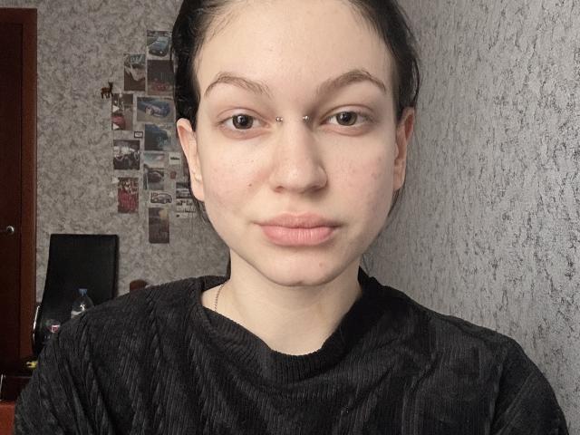 PalmaPrum hat eine helle Haut, dunkle Augenbrauen, eine kleine Nase und volle Lippen. Sie trägt ein schwarzes Hemd, hat ein kleines Nasenpiercing und ihr Haar ist hochgebunden..