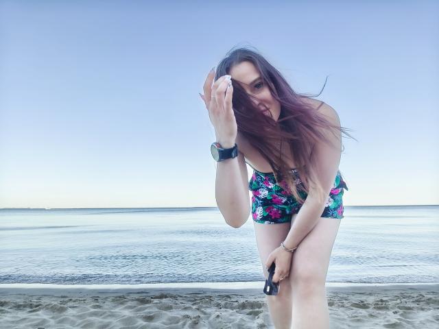 LadyAlessa met lang rood haar, een bloemenbikini, gebogen op het strand, met een telefoon, horloge en armband in haar handen, haar achterkant is licht zichtbaar, een helderblauwe lucht, kalm water, een zandstrand..