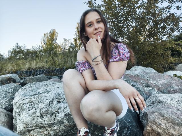 LadyAlessa sentada em pedras, vestindo uma blusa florida com um ombro de fora, shorts brancos e tênis pretos, com o dedo nos lábios, parecendo fofa e brincalhona..