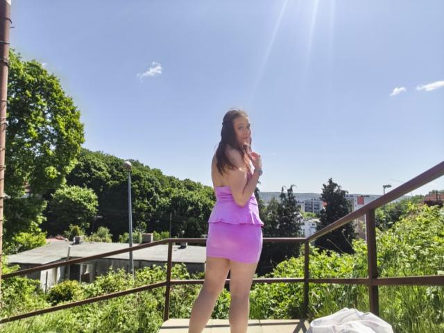 LadyAlessa, con el pelo largo y castaño, vestida con un mini-vestido morado sin tirantes y con volantes, está de pie en un balcón iluminado por el sol, mostrando su trasero desnudo y sujetándose el pecho, mirando hacia atrás..