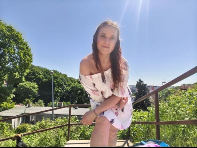 LadyAlessa met lang rood haar, een witte jurk met een open schouderlijn en roze bloemmotieven, zittend op de trap, zonlicht, blauwe lucht, groene bomen en een heldere dag, kijkend iets naar beneden..