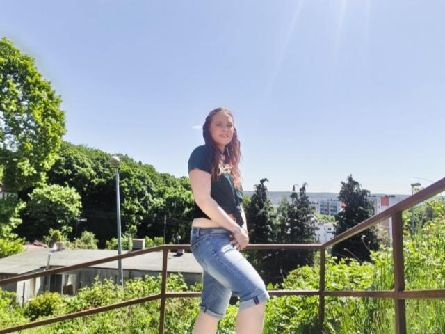 LadyAlessa mit roten Haaren, einem schwarzen T-Shirt und hochgekrempelten blauen Jeans, steht auf einem sonnigen Metallgeländer, die Hände in den Hüften, und blickt in die Kamera. Im Hintergrund sind grüne Bäume und ein klarer Himmel zu sehen..