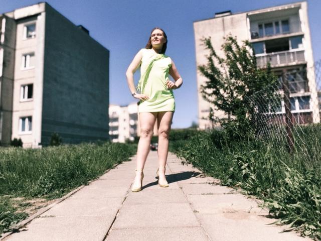 LadyAlessa steht auf einem Bürgersteig, trägt ein neon-grünes Kleid, weiße Schuhe mit Absatz, hat die Hände in die Hüften gestemmt und lächelt, während sie nach unten schaut. Sie ist groß und schlank, der Himmel ist blau und im Hintergrund ist eine städtische Umgebung zu sehen..