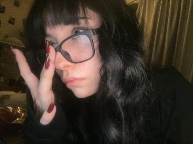 Desiree Rampulla, con cabello negro y ondulado, gafas y uñas rojas, tocándose la cara con una expresión seria, vistiendo una blusa negra, en una habitación con poca luz..