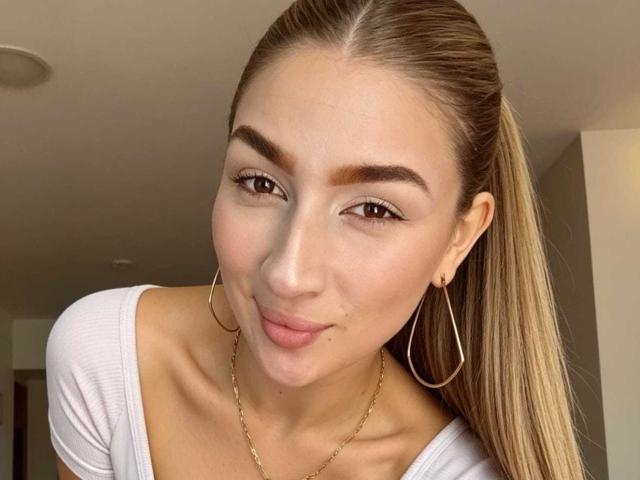 A AbbyMilers tem um sorriso adorável, olhos castanhos e um rabo de cavalo loiro comprido. Ela está vestindo uma blusa branca, brincos de argola dourados e um colar dourado. Seus seios são pequenos e ela é sexy..