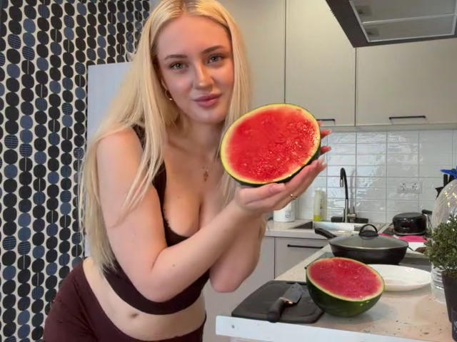 NancyBijou, com cabelo loiro, seios médios em um sutiã preto, segurando metade de uma melancia com polpa vermelha, em pé em uma cozinha, vestindo calças vinho..