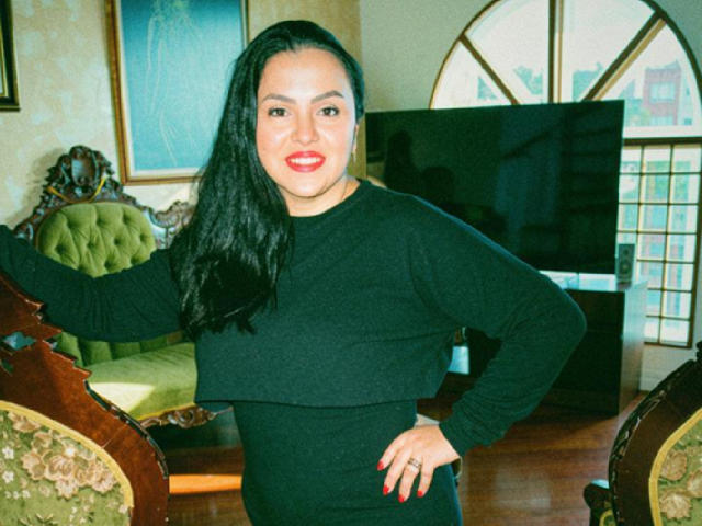 Charlotte Devereaux, con lunghi capelli neri, rossetto rosso e una maglia a maniche lunghe nera, è in piedi con una mano appoggiata a una sedia, mentre guarda verso la fotocamera. Si vede un pavimento di legno e una sedia imbottita di colore verde..