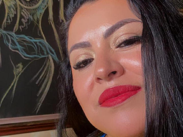 Charlotte Devereaux, met glanzende gouden oogschaduw, dikke zwarte wenkbrauwen en rode lippenstift, lacht met donker zwart haar, een glanzende huid en een lichte frons, en ze ziet er zelfverzekerd uit..