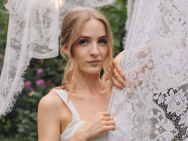 Eva Daly, con i capelli biondi, un abito di pizzo bianco, che tiene una delicata tenda di pizzo, un leggero sorriso, occhi verdi, figura esile, trucco leggero, un anello alla mano destra, luce naturale soffusa..