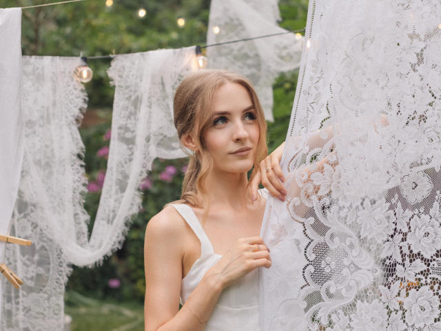 Eva Daly, con piccoli seni, indossa un abito bianco in pizzo, tiene in mano del tessuto in pizzo, è in piedi vicino a delle luci a stringa, guarda verso l'alto con un sorriso appena accennato, in un giardino verde..