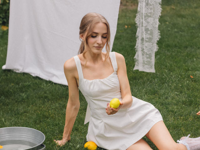 Eva Daly, con i capelli biondi, piccoli seni, vestito bianco, che tiene in mano un limone giallo, seduta sull'erba, con un secchio di metallo pieno di limoni, una tenda di pizzo e un panno bianco sullo sfondo..