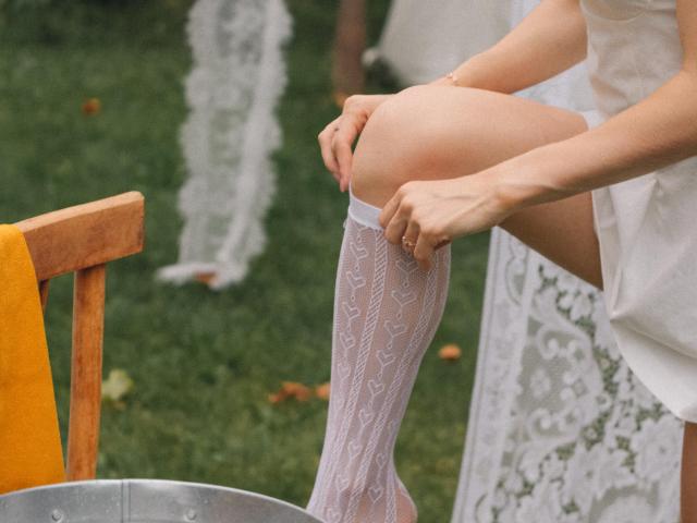 Eva Daly sta indossando calze bianche con un motivo a cuori, tenendosi le ginocchia con le mani. Il suo sedere nudo è parzialmente visibile. Una sedia di legno con un rivestimento giallo è nelle vicinanze..