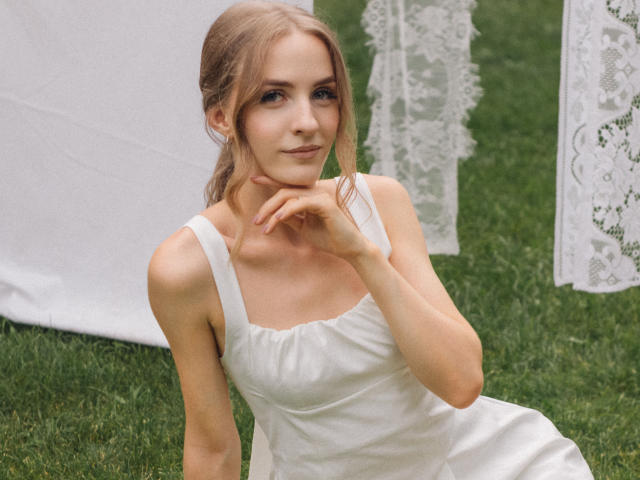 Eva Daly, con i capelli biondi, indossa un abito bianco, seduta sull'erba, con una mano che tocca il mento, seno piccolo, figura snella, carina, sorride dolcemente, tende di pizzo sullo sfondo, erba verde..