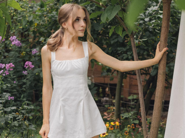 Eva Daly, con piccoli seni, indossa un vestito bianco senza maniche, è in piedi accanto a un albero, che tiene con una mano, e guarda di lato, in un giardino verde..