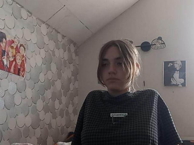 BessBetak, met bruin haar, draagt een zwart geruit shirt en staat in een kamer met een serieuze uitdrukking, waarbij ze iets naar de zijkant kijkt. Kleine zwarte ventilator aan de muur..