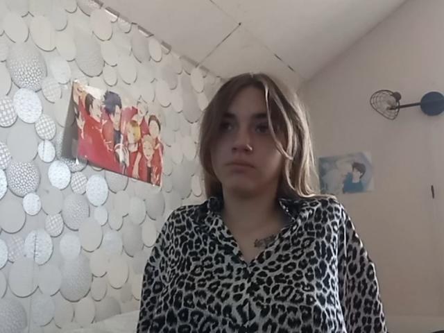 BessBetak, mit braunen Haaren, trägt ein Oberteil mit Leopardenmuster und blickt mit einem neutralen Gesichtsausdruck nach oben. Sie hat ein kleines Tattoo auf ihrer Brust. Ihre Nase ist gestochen..