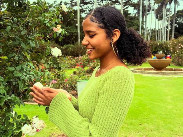 Cinthia Walker, com cabelos escuros e cacheados, vestindo um suéter verde, segurando uma rosa branca, sorrindo, em pé em um jardim, usando brincos grandes em formato de argola, olhando para a rosa, com um fundo de grama, em um dia nublado..