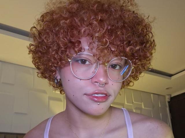 KeiryGinger, com cabelo ruivo encaracolado, óculos, piercing no nariz, piercing no lábio e sardas, vestindo uma camiseta branca, tem um piercing no septo e um colar de ouro..