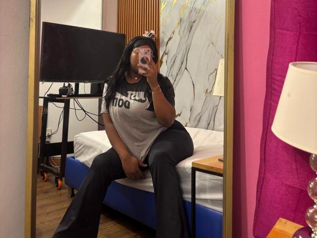 Une jeune femme, DarkTabooGirl, avec de grandes seins, portant un t-shirt gris avec l'inscription "WELCOME" et un pantalon noir, assise sur un lit, prenant un selfie devant un miroir, tenant un téléphone, dans une pièce aux murs roses..
