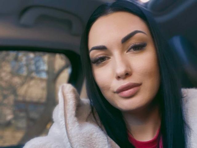 Xioni has dark hair, thick eyebrows, and full lips, wearing a white fluffy jacket, with a slight smile, sitting in a car. Her eyes are half-open, looking forward.