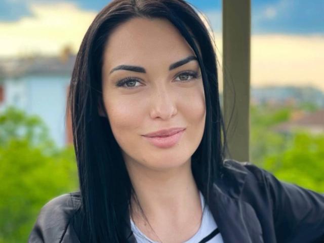 Xioni has black hair, green eyes, and a slight smile with pink lips. She's wearing a black jacket over a white shirt. Her eyebrows are dark and well-defined.