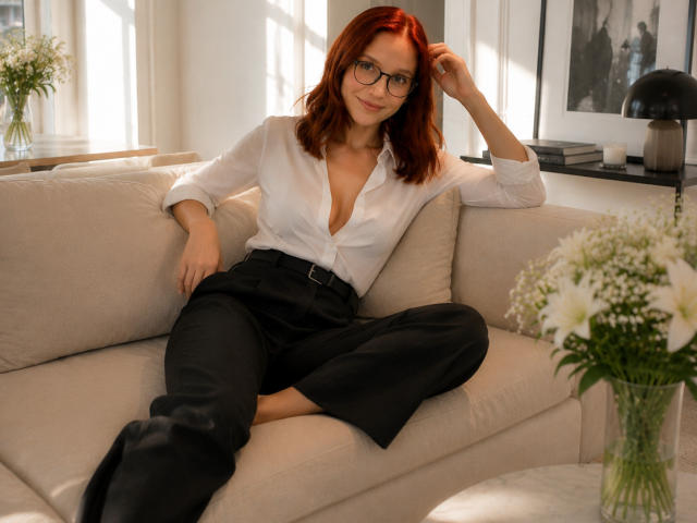 JuliannaMoor with red hair, glasses, white blouse, black pants, sitting on beige couch, hand in hair, slight smile, white flowers in foreground, sunlight, casual, sexy.