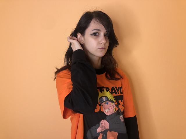 LoyceVannah, con cabello oscuro, una camiseta con diseño negro y naranja, y delineador de ojos grueso, tocándose la cabeza, mirando ligeramente hacia un lado, frente a una pared naranja lisa..