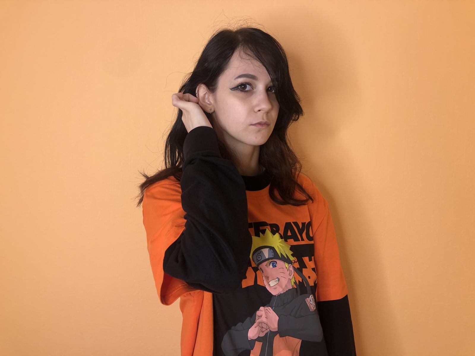 LoyceVannah, avec ses cheveux foncés, son t-shirt graphique noir et orange, et son eye-liner épais, touchant sa tête, regardant légèrement sur le côté, devant un mur orange uni..